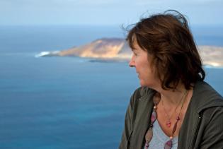 Claudia im Vordergrund schaut von oben auf das unter ihr liegende Meer. Im Hintergrund die kleine Insel La Graciosa.