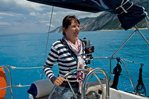 Claudia am Steuerrad der Segelyacht, im Hintergrund die helle Felsküste der Insel Lefkáda