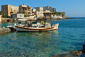 Im Vordergrund ein farbiges Fischerboot angeleint am Hafenrand; im Hintergrund steinernde Häuser rund um das Hafenbecken.